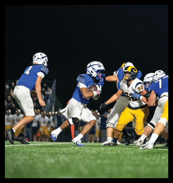 Two people in football uniforms are engaged in a play on a field at night with one person holding the ball and the other reaching out to block or tackle. Description generated by AI