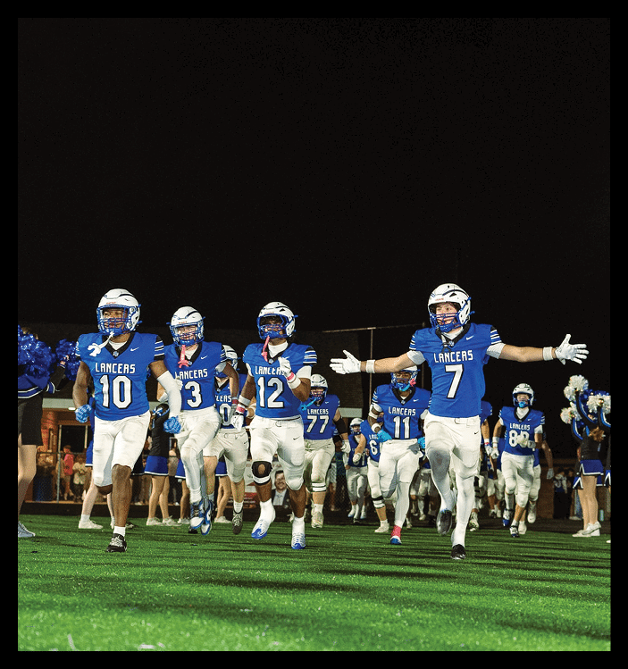 A group of people wearing blue and white football uniforms with “Lancers" on their jerseys are running energetically onto a green field at night with cheerleaders visible in the background. Description generated by AI