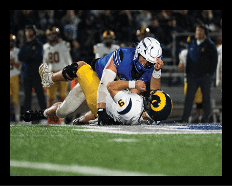 Two people in football uniforms are engaged in a tackle on a field during a game, with one person in a blue jersey pinning another in a white jersey with a yellow helmet to the ground. Description generated by AI