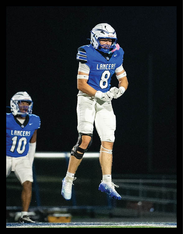 Two people wearing blue football uniforms with white helmets and pants are on a field at night, with one person standing and clenching their fists while the other is slightly crouched. Description generated by AI
