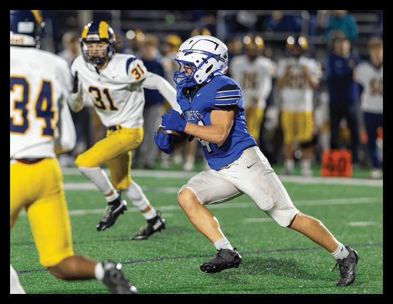 A person in a blue football uniform is running with the ball while another person in a white and yellow uniform chases behind on a football field. Description generated by AI