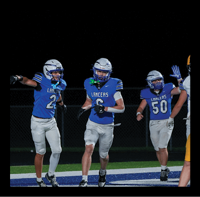 Two people in blue football uniforms and white helmets are on a field at night, with one person pointing and the other holding a mouthguard. Description generated by AI