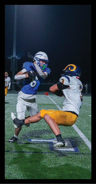 Two people in football uniforms are engaged in a play on a lit field at night, with one person in a blue jersey preparing to throw the ball and the other in a white and yellow uniform attempting to block or tackle. Description generated by AI