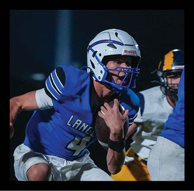 Two people in blue and white football uniforms are actively playing on a field at night with another person in a yellow and white uniform behind them. Description generated by AI