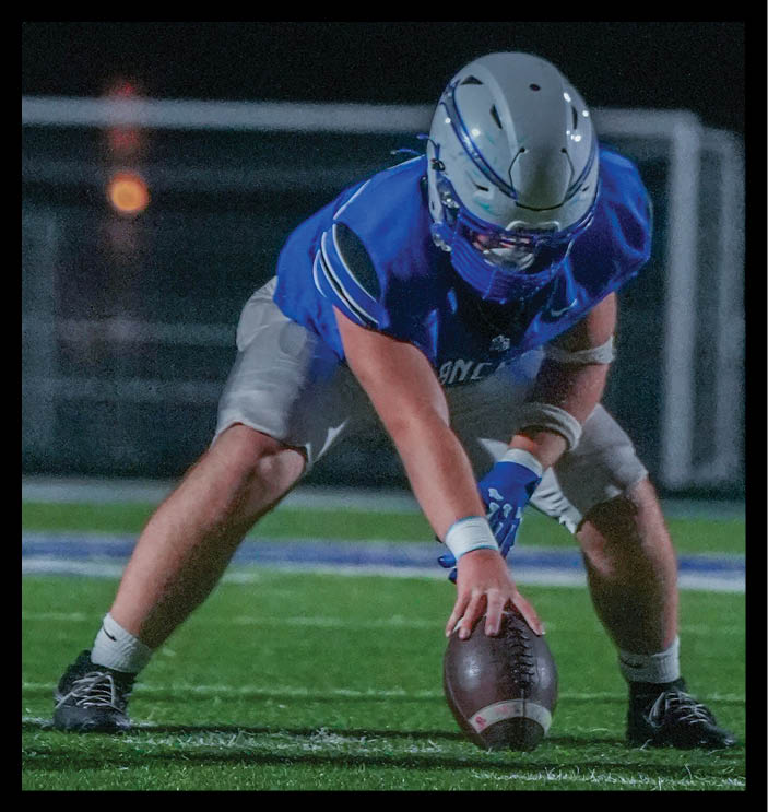 Three people in football uniforms are positioned on a field at night, with one person holding a football ready to snap it and another standing behind, appearing to prepare for a play. Description generated by AI