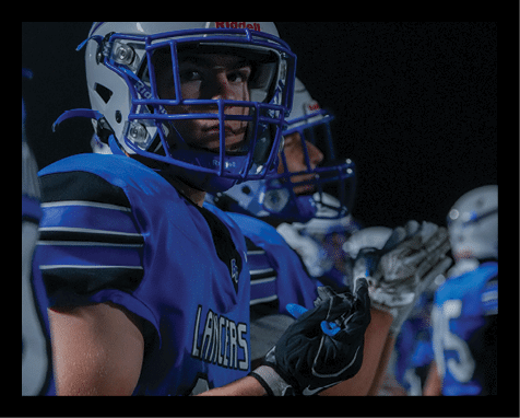 A person in a blue football uniform and helmet is looking toward the camera while standing next to other similarly dressed people in a dark outdoor setting. Description generated by AI