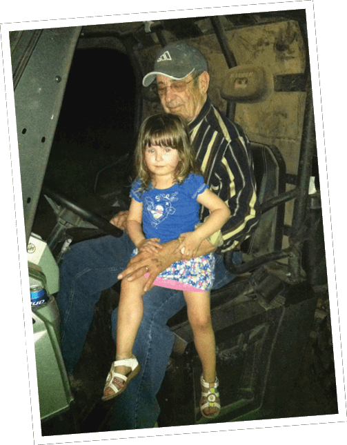A senior wearing a striped shirt and cap is sitting in a vehicle with a child in a blue shirt and floral skirt sitting on their lap. Descripci n generada con IA