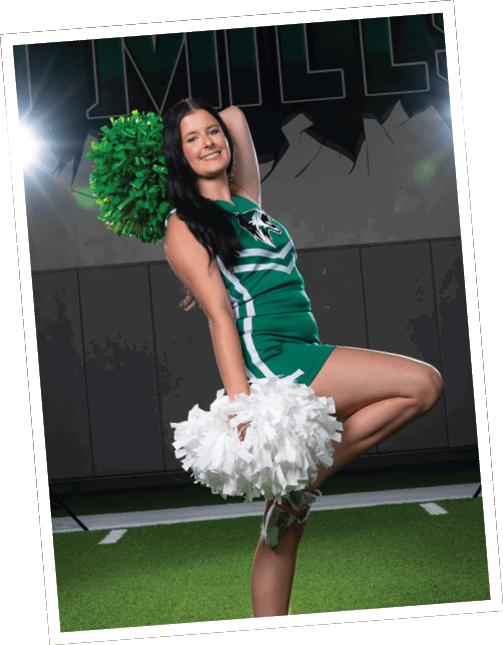 A person in a green and white cheerleading uniform poses on one leg holding green and white pom-poms on a turf field with bright lights and a “MILLS" sign in the background. Descripci n generada con IA