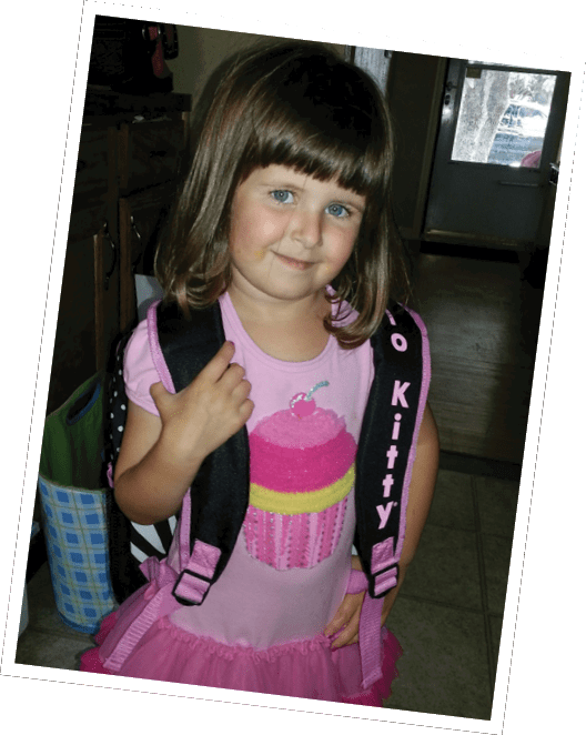 A child with shoulder-length hair wearing a pink shirt with a cupcake design and a pink and black backpack stands indoors near a door and wooden cabinets. Descripci n generada con IA