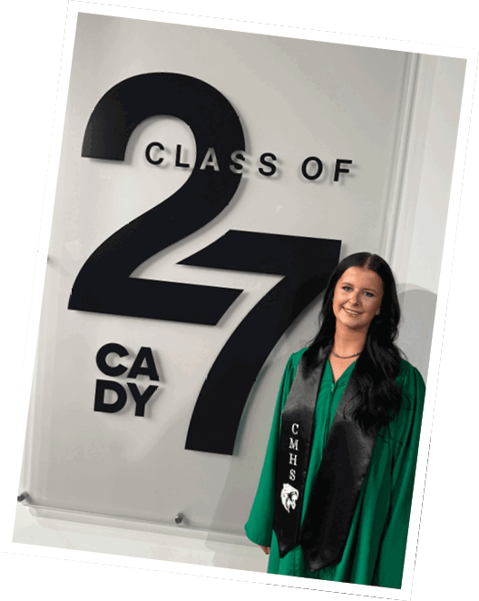 A person with long dark hair wearing a green graduation gown and black stole stands smiling next to a large sign that reads “Class of 27 CADY." Descripci n generada con IA