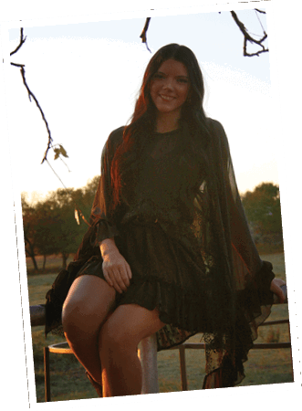 A person with long hair wearing a dark, flowing dress is sitting outdoors on a railing with trees and a sunset in the background. Descripci n generada con IA