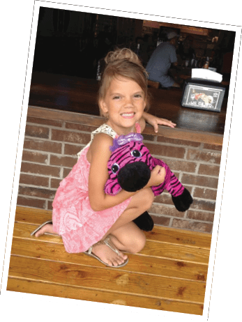 A child with hair tied up is smiling while crouching on a wooden floor and hugging a large pink and black striped stuffed animal with a purple bow. Descripci n generada con IA