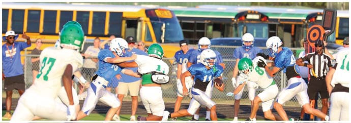 People wearing helmets and sports uniforms are engaged in a football game on a field with school buses and spectators in the background. Descripci n generada con IA