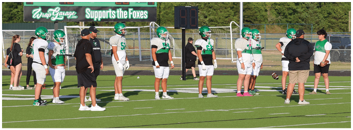 Adults and children wearing green and white football uniforms stand on a field near a scoreboard that reads “Supports the Foxes." Descripci n generada con IA
