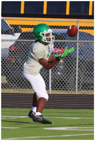 A person wearing a green helmet and white sports uniform is preparing to catch a football on a grassy field near a chain-link fence with parked vehicles and a school bus in the background. Descripci n generada con IA