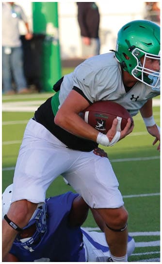 A person in white sportswear is holding a football while another person in blue sportswear is on the ground behind them on a football field. Descripci n generada con IA