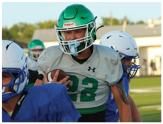 Three people wearing football helmets and uniforms are actively engaged in a play on a football field during daylight. Descripci n generada con IA
