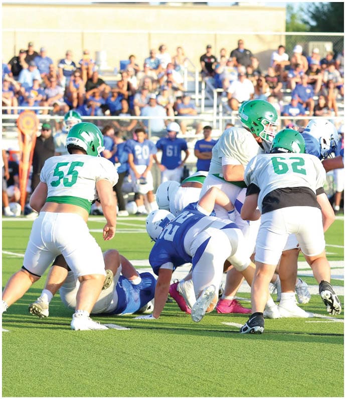 Players in green and white uniforms and players in blue and white uniforms are engaged in a football game on a grassy field with spectators in the background. Descripci n generada con IA