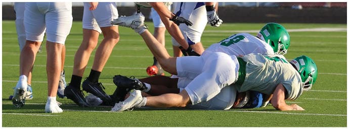 Two people wearing green helmets and gray sports uniforms are tackling a person in a blue uniform on a football field while others stand nearby. Descripci n generada con IA