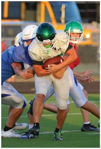 Several people wearing football helmets and uniforms are actively engaged in a tackle on a green football field with a school bus and bleachers in the background. Descripci n generada con IA