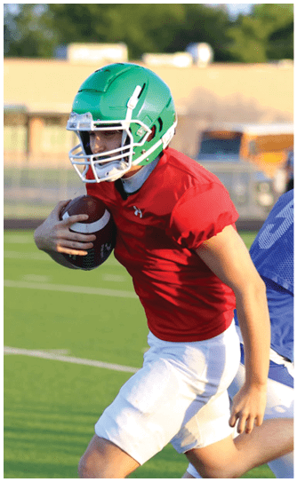 A person wearing a green helmet and red shirt is running with a football while another person in a blue jersey attempts to catch them on a sports field. Descripci n generada con IA