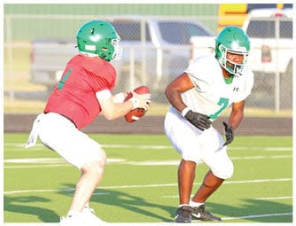 Two adults wearing green helmets and football uniforms are on a football field, with one holding a football and the other positioned in front. Descripci n generada con IA