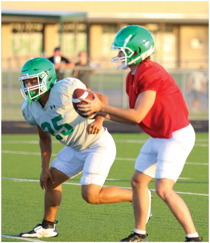 Two people in sports uniforms are playing football on a field, with one person holding the ball and the other reaching to grab it. Descripci n generada con IA