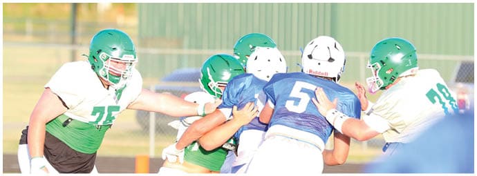 People wearing helmets and sports uniforms are engaged in a football play on a field near a green building. Descripci n generada con IA