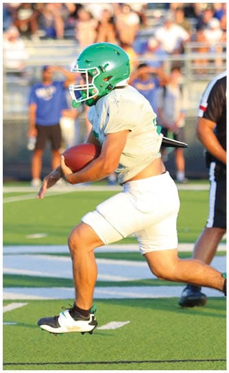 An adult wearing a green helmet and white sports uniform is running with a football on a field while a referee and other players in blue uniforms are visible in the background. Descripci n generada con IA