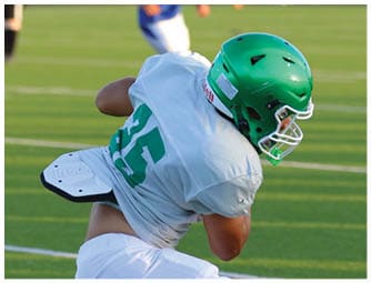 A person wearing a green helmet and white sports jersey with the number 25 is crouched on a grassy field with blurred figures in the background. Descripci n generada con IA