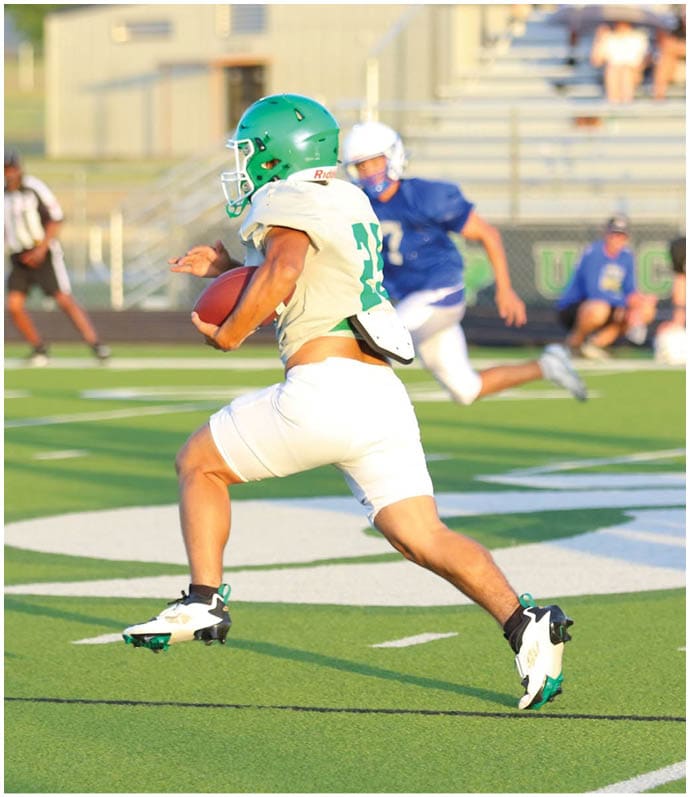 An adult wearing a green helmet and white sports uniform is running on a football field while holding a football. Descripci n generada con IA