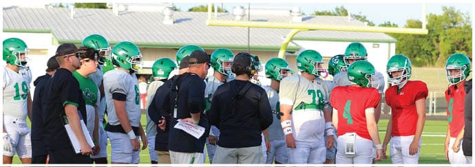 People wearing green and red football helmets and uniforms stand on a field while coaches in black shirts and shorts give instructions. Descripci n generada con IA