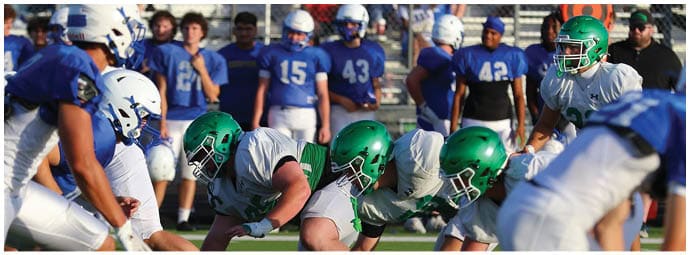 Two opposing American football teams in helmets and uniforms are lined up facing each other on a field with spectators in the background. Descripci n generada con IA