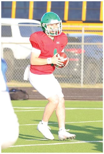 An adult wearing a red jersey and green helmet is holding a football while other adults in white jerseys and green helmets are positioned on a football field with a school bus and fence in the background. Descripci n generada con IA