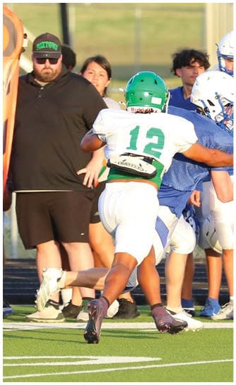 Two people in football uniforms are engaged in a play on a field while a group of people in blue shirts watch from the sidelines near a marker indicating yardage. Descripci n generada con IA