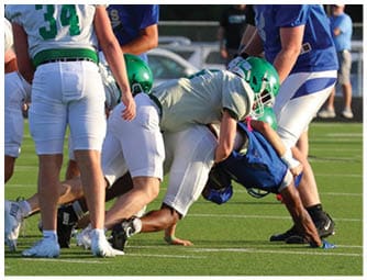 Several people wearing sports uniforms and helmets are engaged in a football tackle on a green field with white yard lines. Descripci n generada con IA