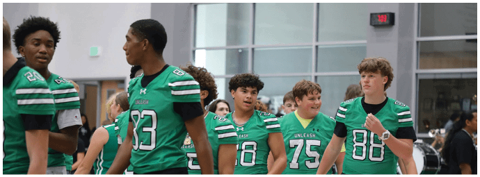 Adults and children wearing green and white football uniforms stand on a field near a scoreboard that reads “Supports the Foxes." Descripci n generada con IA
