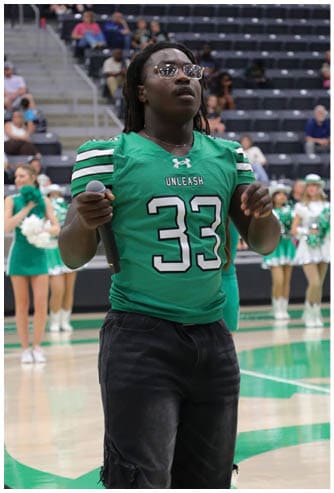 A person wearing a green helmet and white sports uniform is preparing to catch a football on a grassy field near a chain-link fence with parked vehicles and a school bus in the background. Descripci n generada con IA