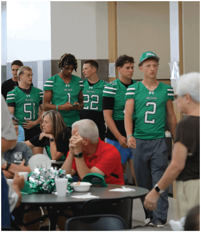 Players in green and white uniforms and players in blue and white uniforms are engaged in a football game on a grassy field with spectators in the background. Descripci n generada con IA