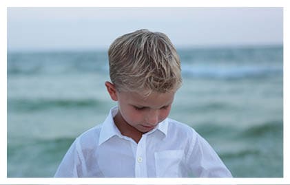 A child with short, light-colored hair is looking down with a blurred ocean and sky in the background. Descripci n generada con IA