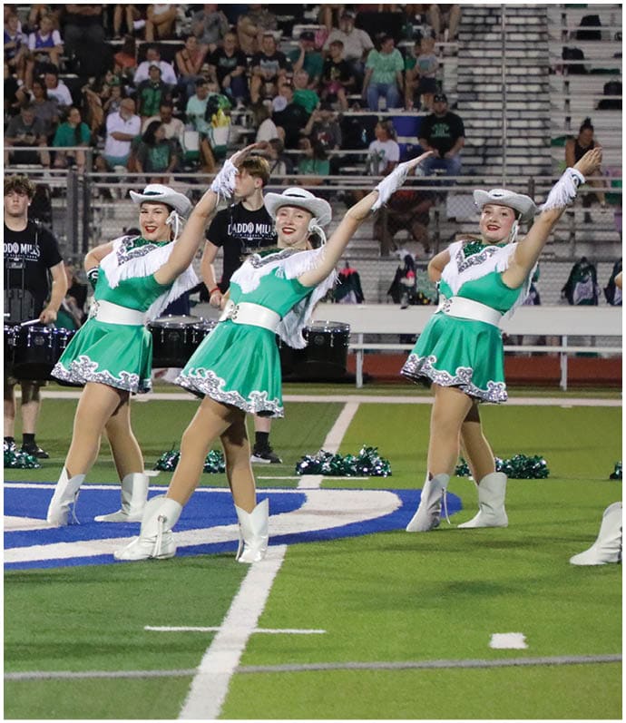 Two adults in green and white dance uniforms with hats perform a synchronized dance on a football field with a marching band and spectators in the background. Descripci n generada con IA