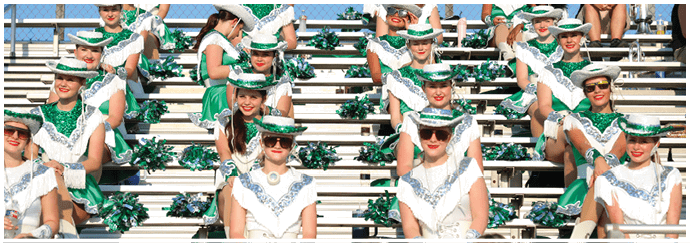 A group of people dressed in coordinated green and white outfits with hats and sunglasses are seated in bleachers holding green pom-poms, likely part of a cheer or dance team. Descripci n generada con IA