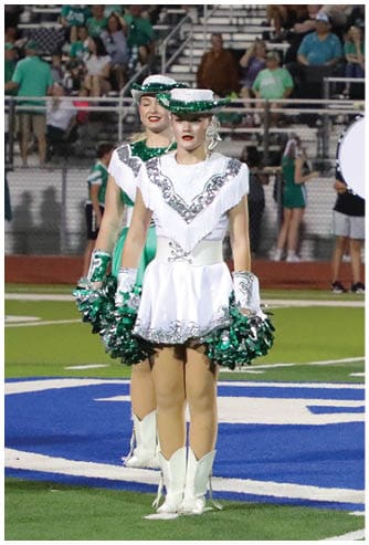 A person in a white sequined uniform with a hat and white boots stands on a football field holding green and silver pom-poms while people in the background play drums. Descripci n generada con IA