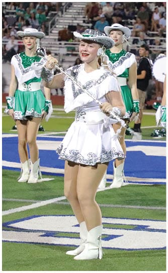 Adults in green and white dance uniforms with hats stand on a football field with a crowd in the background. Descripci n generada con IA