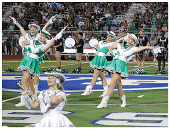 Adults in green and white dance uniforms perform a choreographed routine on a football field with a crowd and a drumline in the background. Descripci n generada con IA