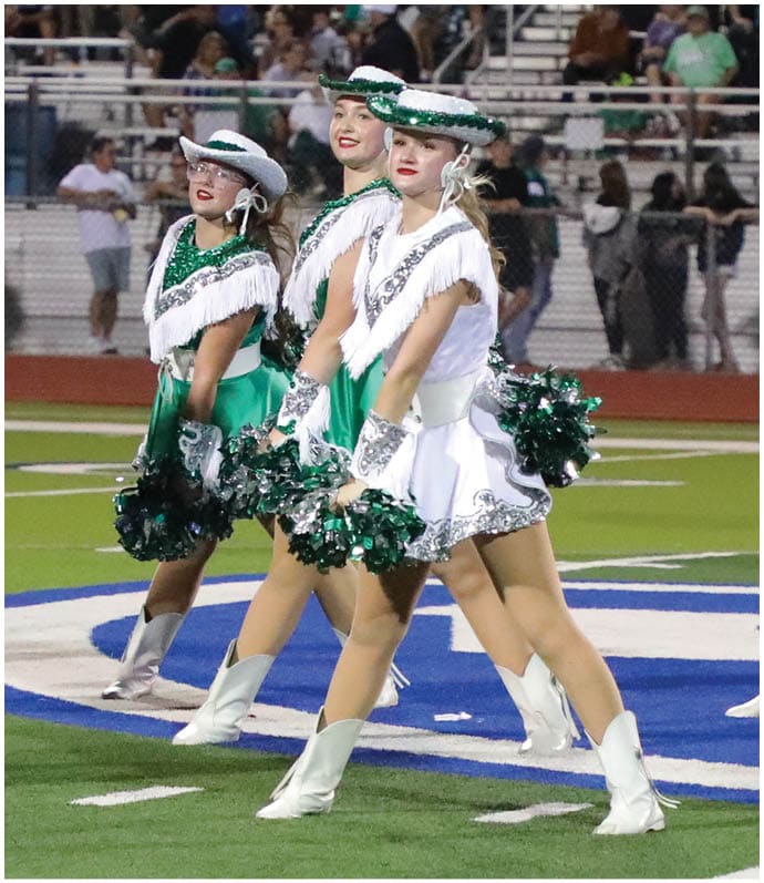 A group of people dressed in white and green dance uniforms with hats and pom-poms perform a coordinated routine on a football field in front of a seated crowd. Descripci n generada con IA