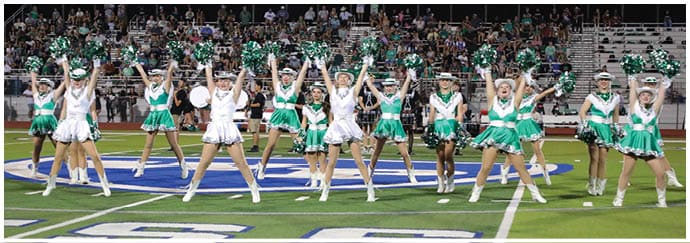 A group of people in green and white cheerleading uniforms perform a synchronized routine on a sports field with a crowd seated in the background. Descripci n generada con IA