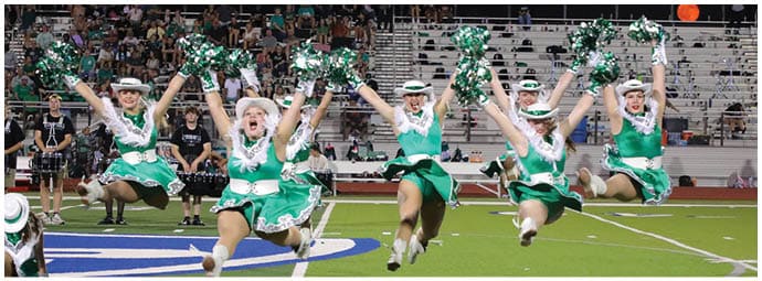 Five people wearing green and white cheerleading uniforms and hats are jumping in unison on a sports field with pom-poms raised above their heads. Descripci n generada con IA