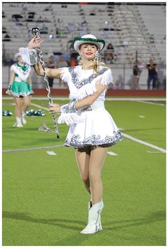A person dressed in a white and green fringed dance costume and hat is performing on a sports field while holding a baton. Descripci n generada con IA
