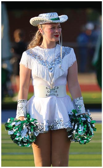 A person wearing a white sequined dance costume with a wide belt, white boots, and a hat is standing on a sports field holding green and silver pom-poms. Descripci n generada con IA